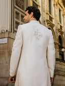 Man wearing a white sherwani with intricate embroidery in front of an ornate building.