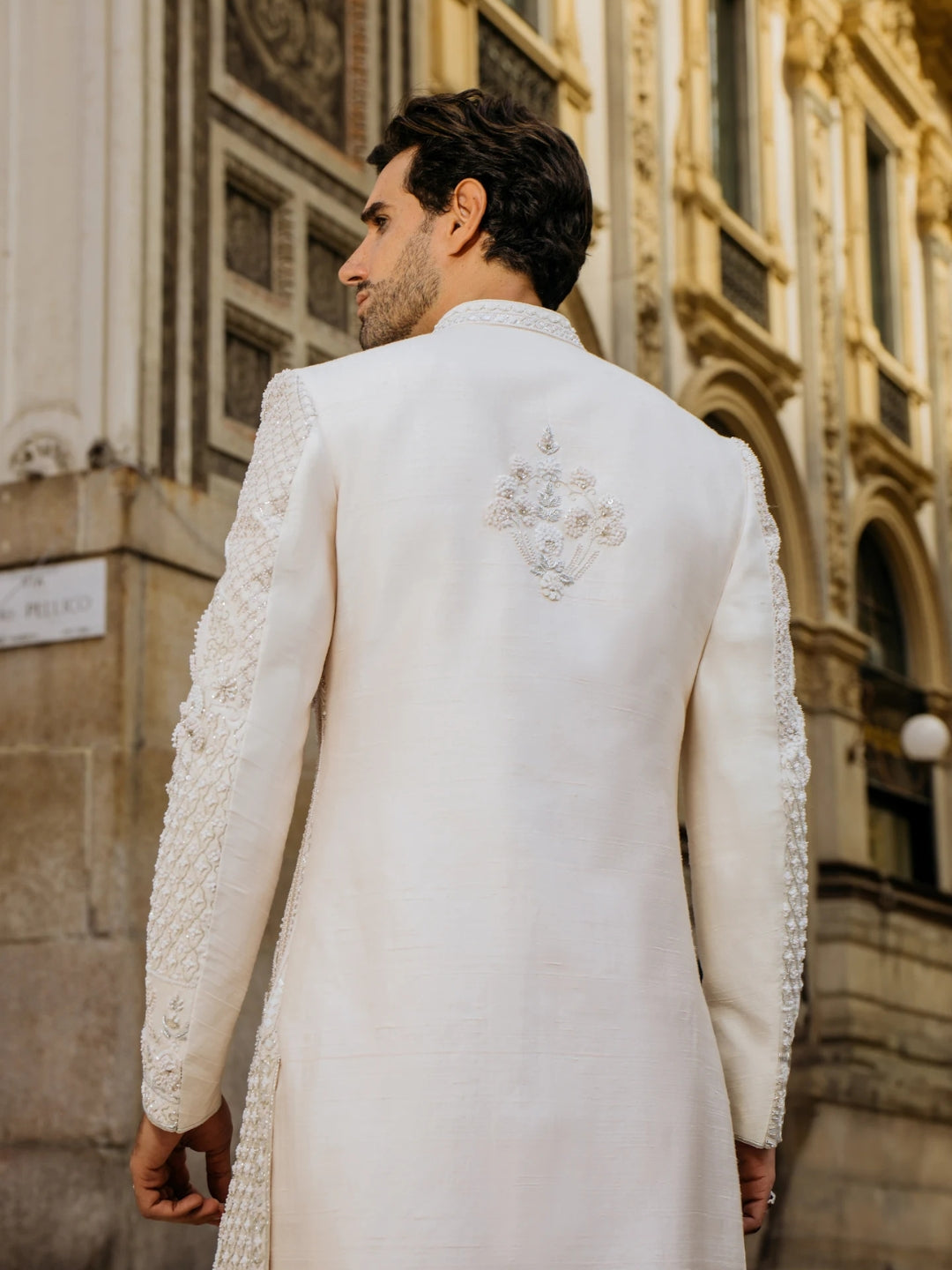 Man wearing a white sherwani with intricate embroidery in front of an ornate building.