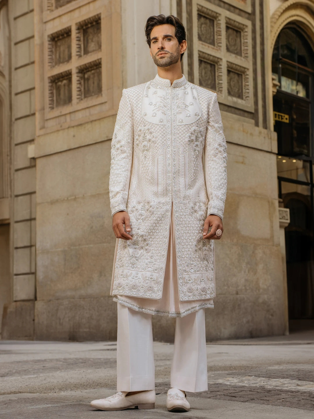 Man wearing a traditional white sherwani standing in front of an architectural building.