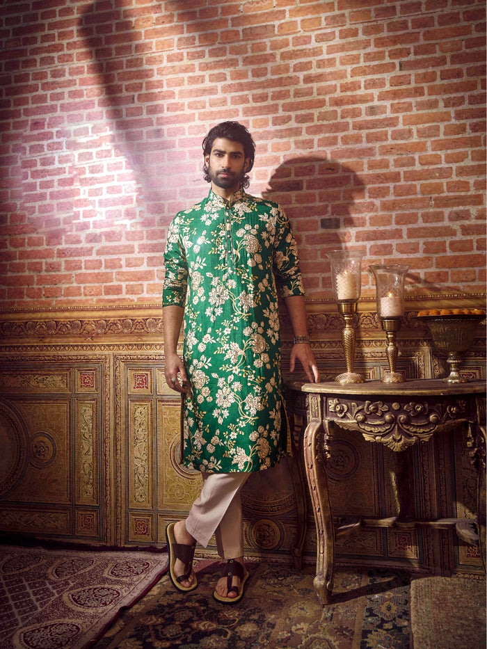 Man in a green floral kurta standing in a room with a brick wall and ornate furniture.